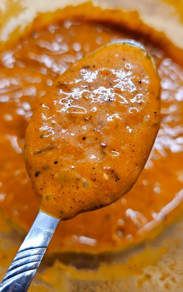 Close-up of a spoonful of thick, orange-brown sauce with visible spices, held above a bowl filled with the same sauce.