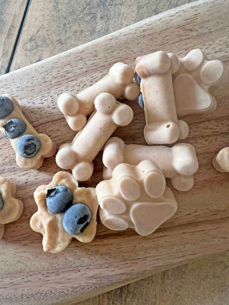 A wooden board with dog-shaped ice cream treats, some adorned with blueberries, placed on a wooden surface.