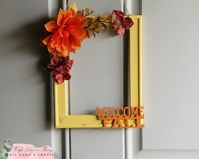 A rectangular yellow picture frame with orange and red flowers is hanging on a door. The frame has the text "WELCOME FALL" in orange letters at the bottom.