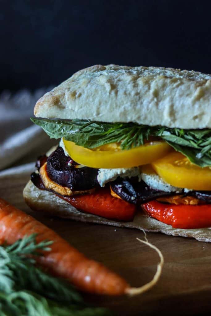 A sandwich with vegetables, including red peppers, yellow tomatoes, leafy greens, and goat cheese, on ciabatta bread. A carrot with greens is partially visible in the foreground.