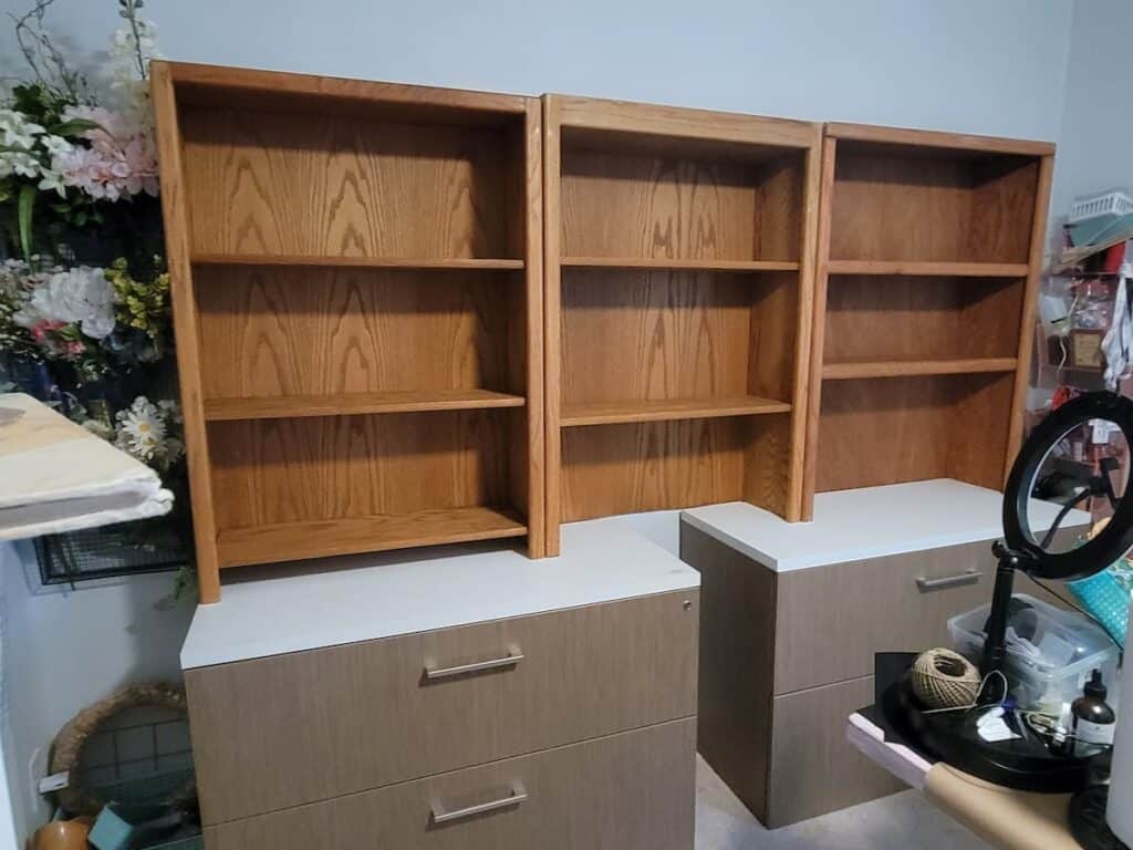 Three wooden bookshelves without items are placed on top of three matching two-drawer filing cabinets in a cluttered room with various items and a ring light nearby.