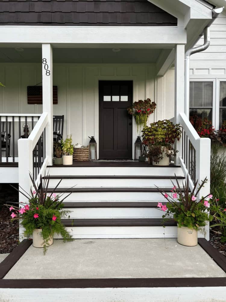 A neatly kept front porch with potted plants on either side of the steps, a dark front door, house number 808 on a white post, and a small American flag hanging on the wall.