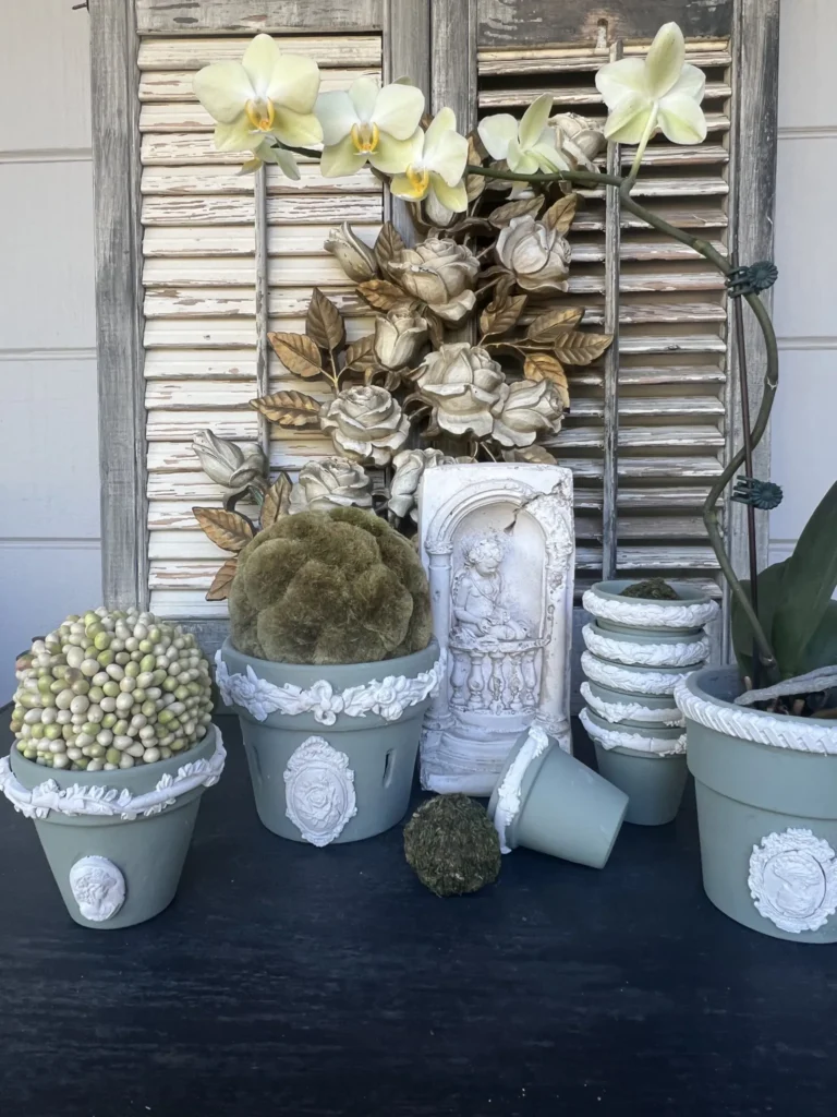 A collection of decorative pots with various plants, including a potted orchid with white flowers, displayed in front of distressed wooden shutters.