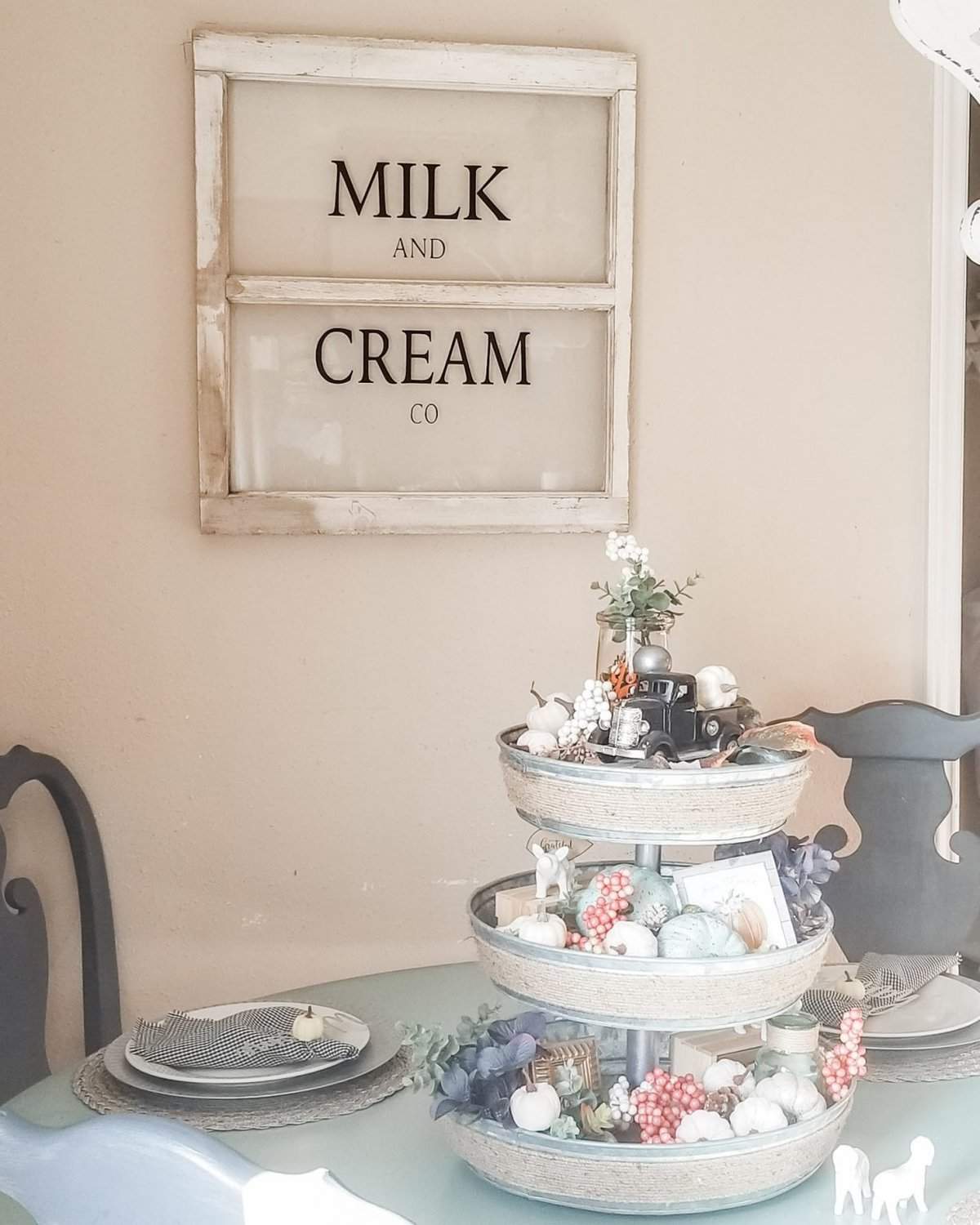 A tiered tray with assorted decorations sits on a round table with place settings. Behind the table, a wall sign reads "Milk and Cream Co." in a vintage window frame.