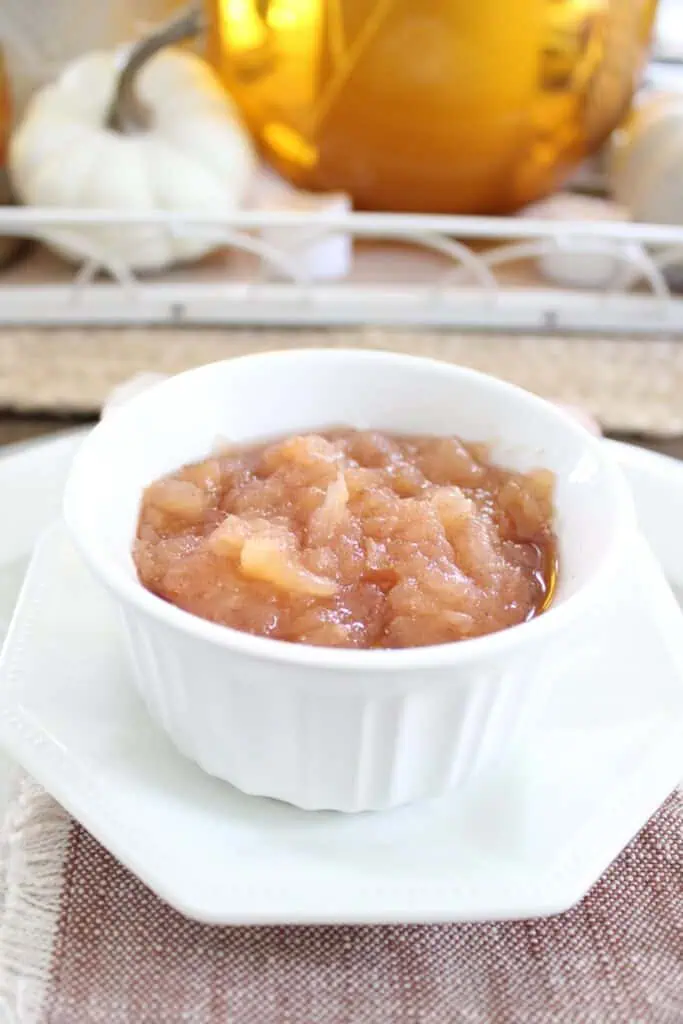 A white bowl filled with chunky applesauce sits on a matching plate with a brown fabric underneath. In the background, there are decorative items including a white pumpkin and a jar.