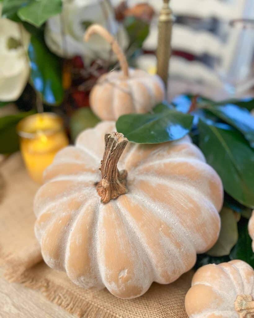 Close-up of a pale, ribbed pumpkin with a rough stem, surrounded by green leaves and a small, yellow candle in the background.