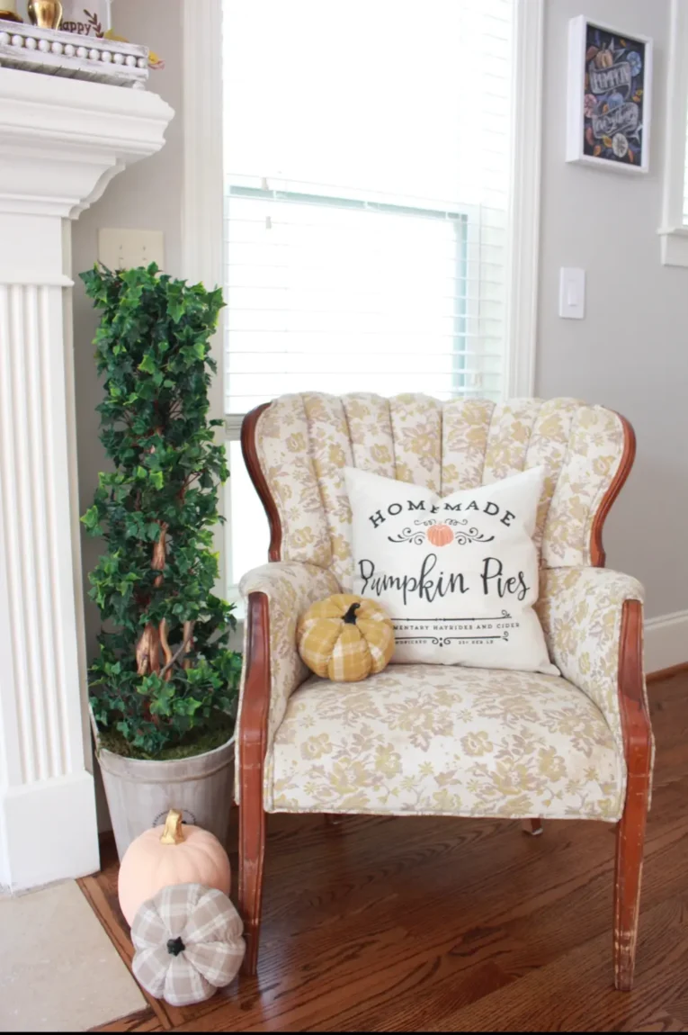 A decorative chair with a floral pattern holds a cushion that reads "Homemade Pumpkin Pies." In front of the chair are two fabric pumpkins, and beside it stands a potted artificial plant.