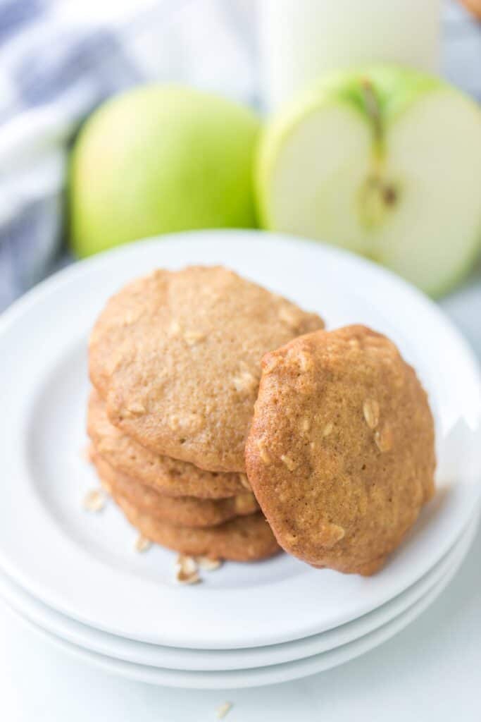 A white plate with a stack of cookies is placed in front of a halved green apple and a whole green apple, along with a blurred blue and white checkered cloth in the background.