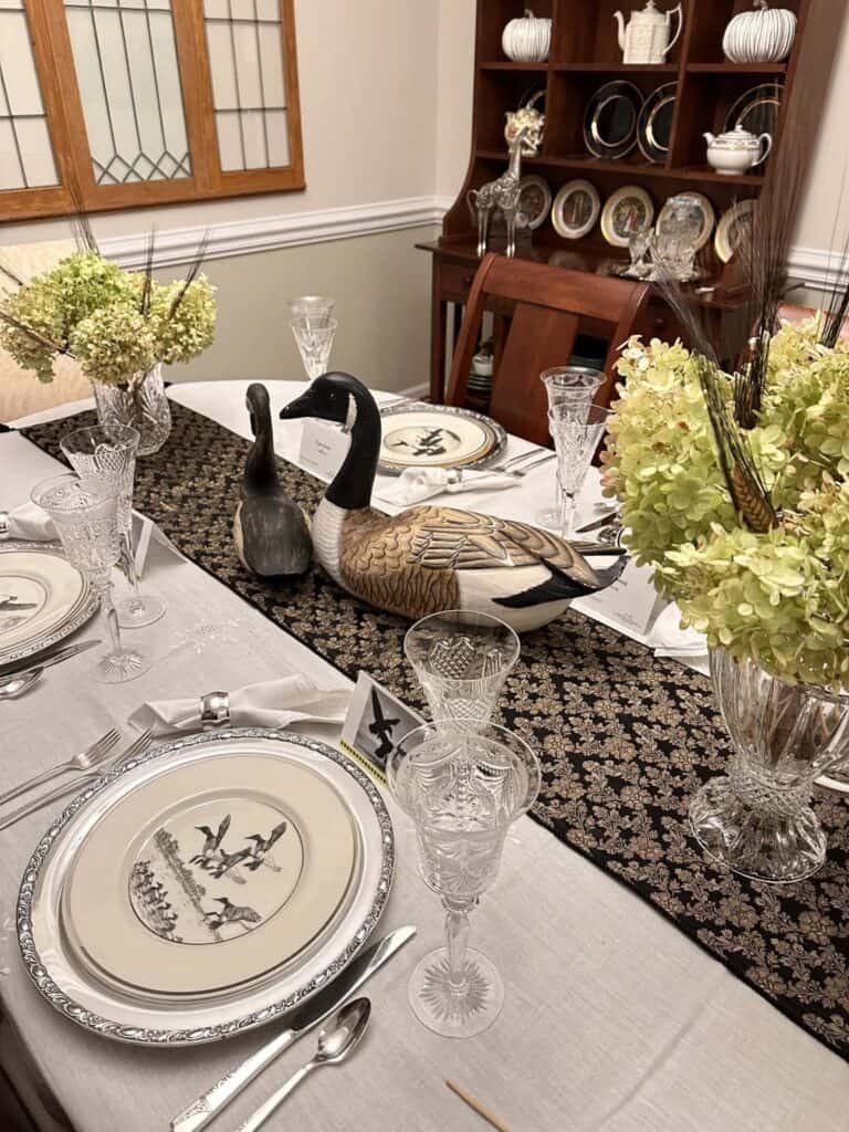 A dining table set with crystal glassware, patterned plates, and a decorative black table runner featuring two wooden duck sculptures. The background shows a hutch with china plates and glassware.