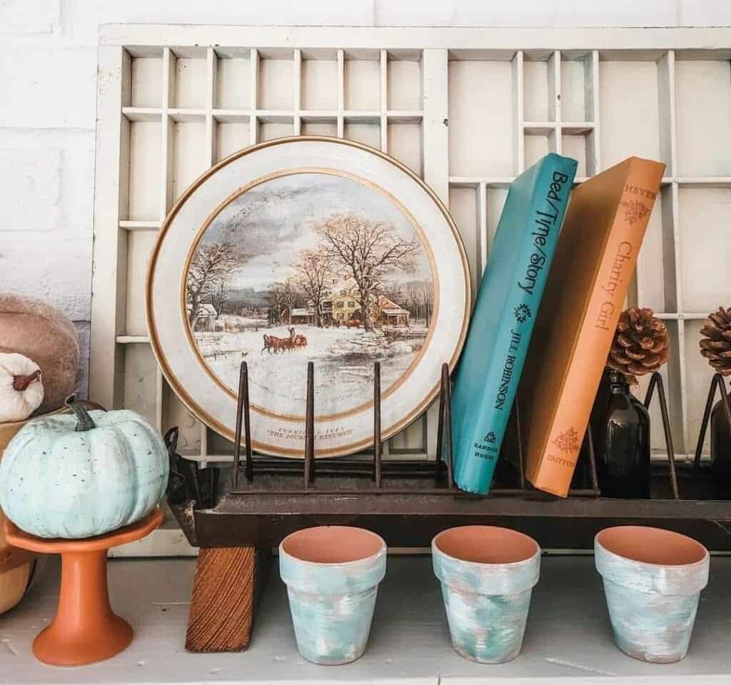 Shelf display with a winter scene plate, two books, a small blue pumpkin on a stand, and three painted terra cotta pots. Pine cones and a metal tray add rustic accents.