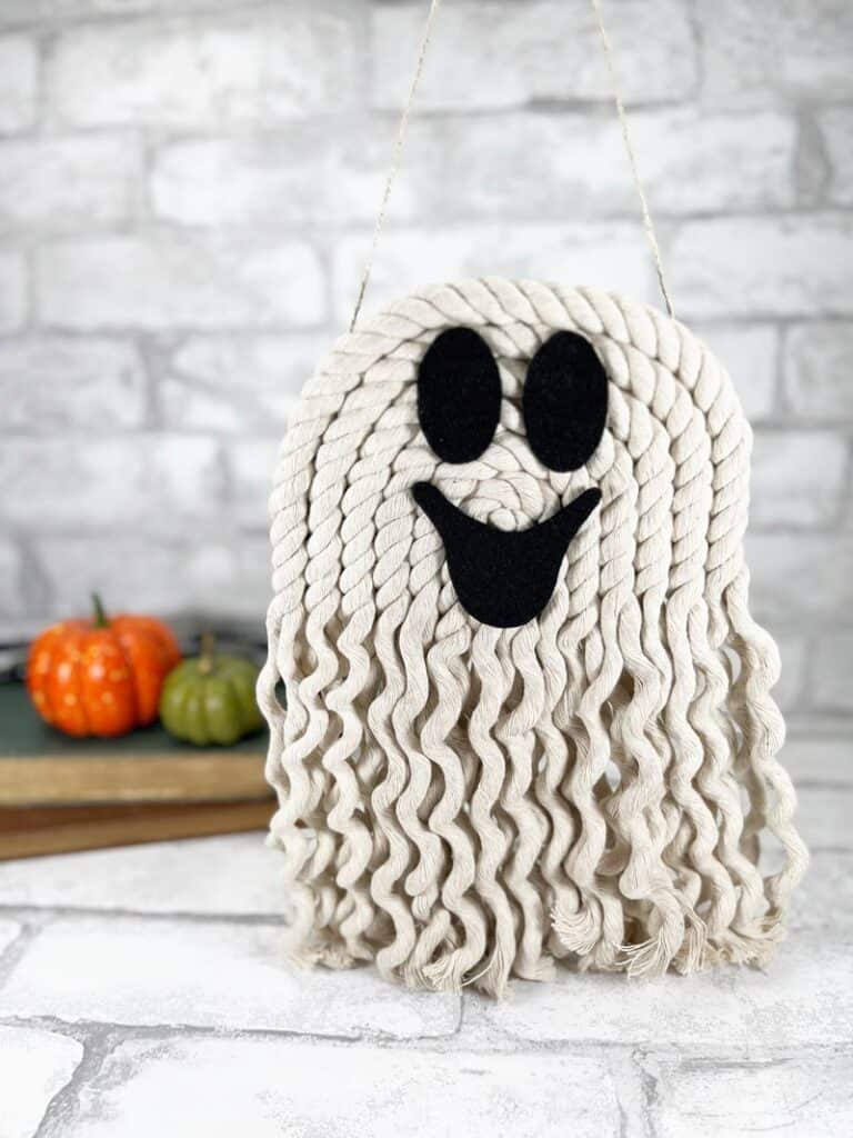 Braided yarn ghost decoration with black felt eyes and mouth, hanging against a brick backdrop. Small pumpkins and books are in the background.