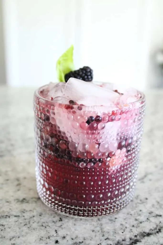 A textured glass filled with a pink and red berry drink, topped with ice, blackberries, and a mint leaf.