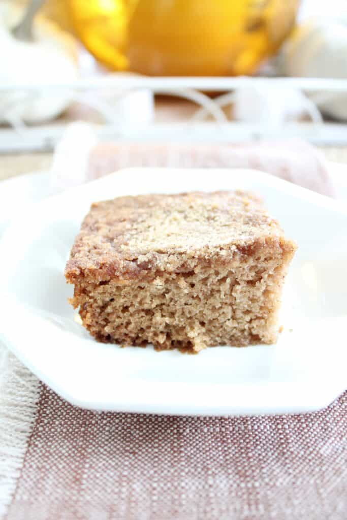 A square piece of cinnamon coffee cake on a white plate, resting on a cloth napkin.