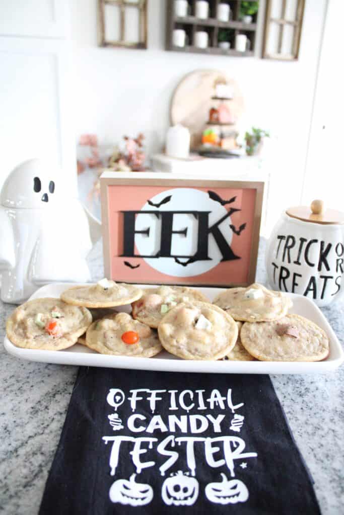 A plate of Halloween cookies on a counter, surrounded by decor including a ghost jar, a "Trick or Treat" container, and a sign reading "EEK" with bats. A "Official Candy Tester" napkin lies in front.