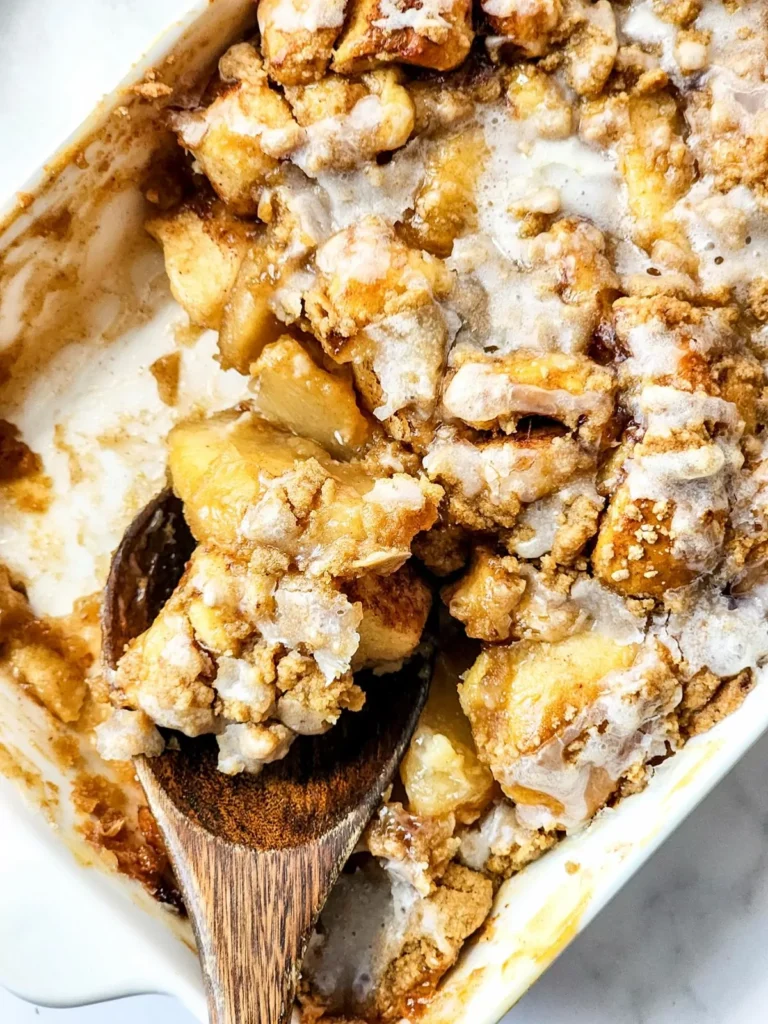 A close-up of a baked apple crumble in a dish with a wooden spoon. The crumble topping is golden brown and looks crispy and bubbled with juices.