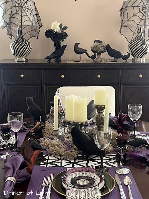 A dining table with a Halloween theme, featuring candles, raven figurines, and spider web decorations. Glassware and cutlery are neatly arranged.