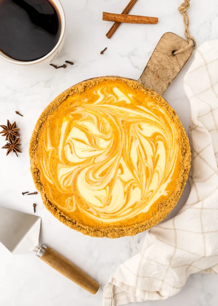 A pumpkin swirl pie on a wooden board next to a cup of coffee, a pie server, and a white cloth, with cinnamon sticks and star anise nearby.