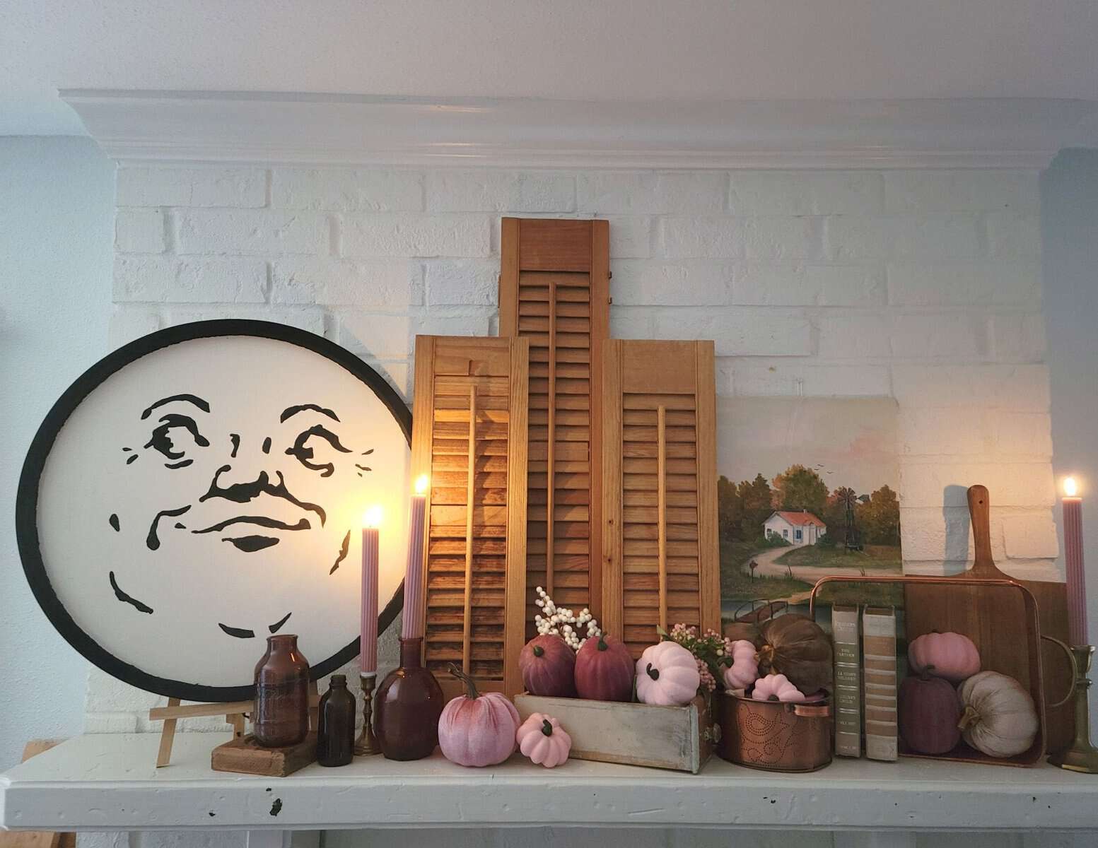 A mantelpiece display with a round face illustration, wooden shutters, pumpkins, candles, and a landscape painting against a white brick wall.