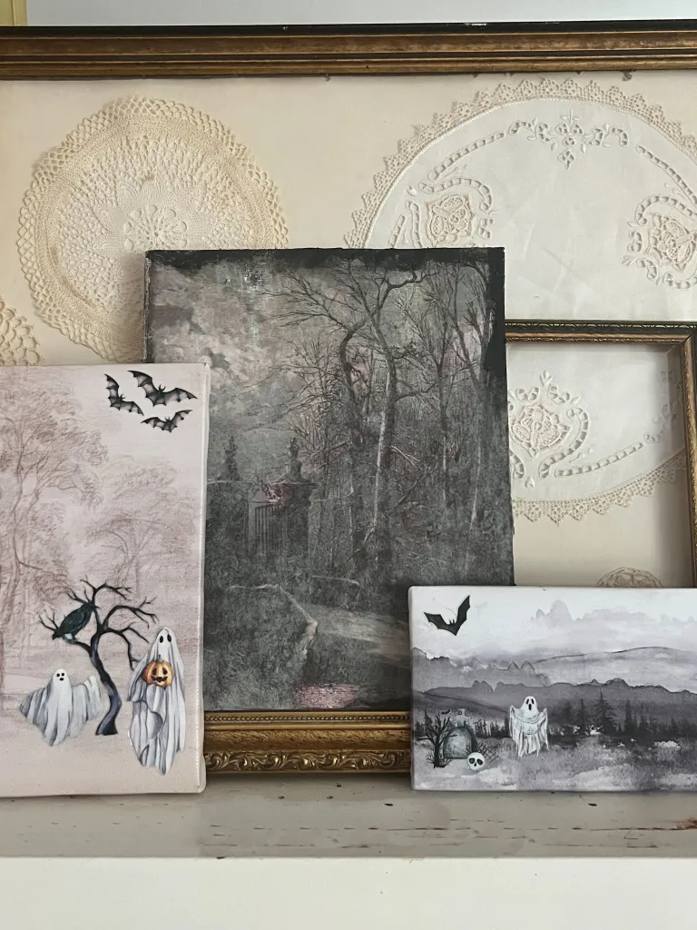 Three framed artworks featuring ghostly themes, with one showing trees, another with flying bats, and the third displaying a misty landscape. Background has an embroidered lace design.