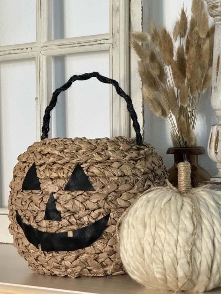 A woven basket designed as a jack-o'-lantern sits on a shelf beside a white pumpkin and a vase with dried grasses.