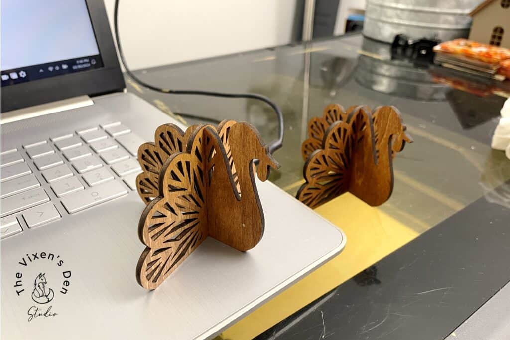 Two wooden turkey-shaped decorations sit beside a laptop on a reflective surface, next to a logo that reads "The Vixen's Den Studio.