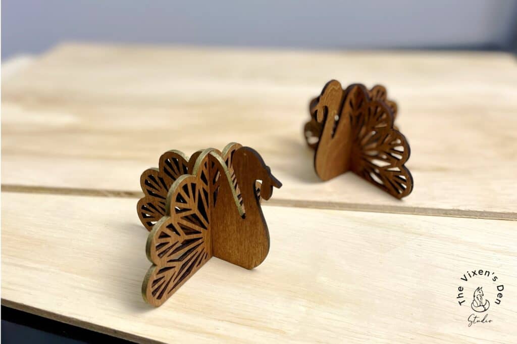 Two wooden turkey-shaped decorations with intricate cut-out details are positioned on a light wooden surface. The logo "The Vixen's Den" is visible in the corner.