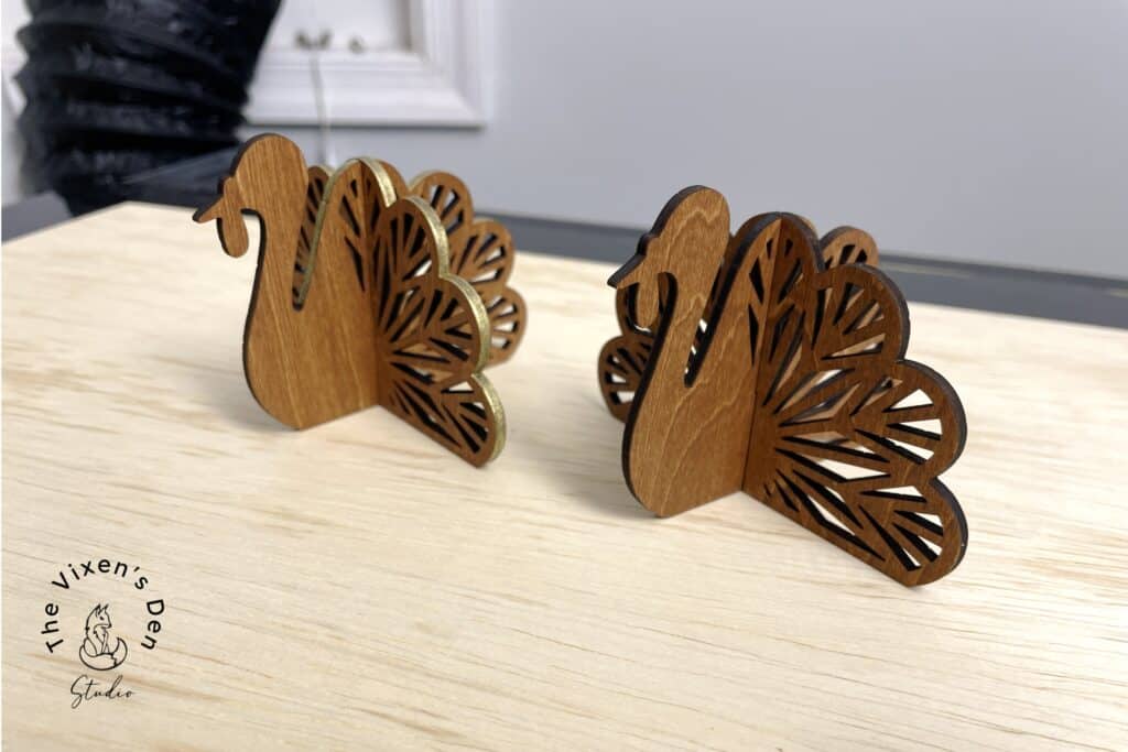 Two wooden peacock-shaped decor pieces on a light wooden surface with a logo in the corner.