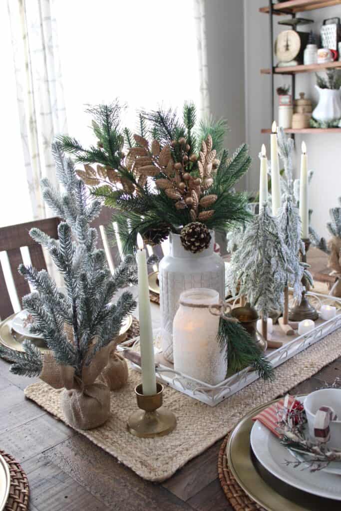 A festive dining table with candle holders, small Christmas trees, and a central arrangement of pine branches and gold accents. Plates and napkins are neatly arranged around the table.