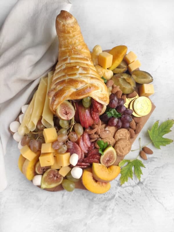 A cornucopia-shaped pastry filled with assorted fruits, cheese cubes, nuts, and cookies on a wooden board, with a white cloth on the side.