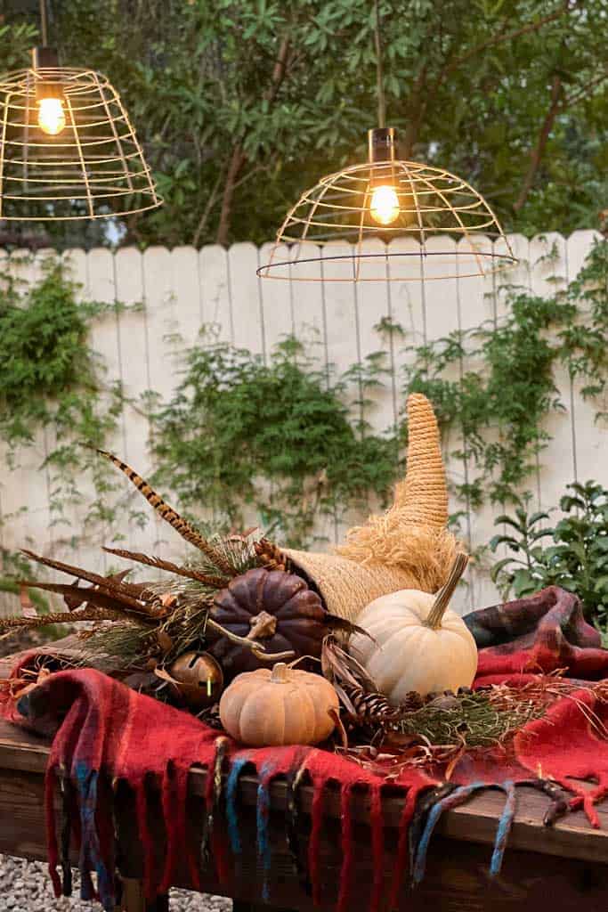 Table with pumpkins, cornucopia, and fall decor on a red blanket in an outdoor setting with two wire pendant lights above and greenery on a white fence backdrop.