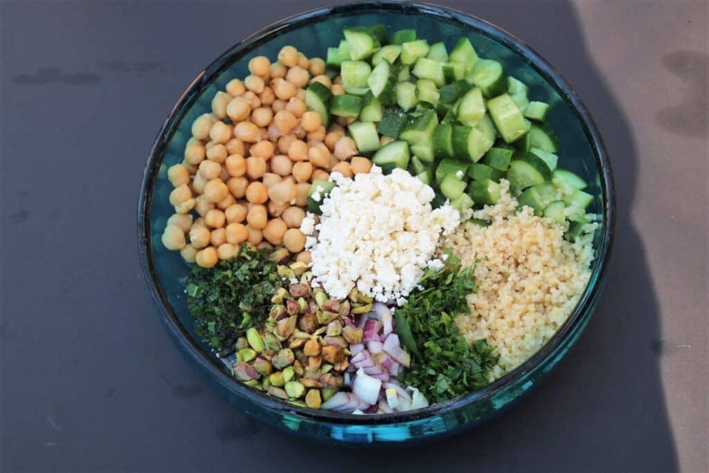 A bowl with chickpeas, diced cucumber, crumbled feta, bulgur, chopped mint, red onion, pistachios, and parsley on a dark surface.