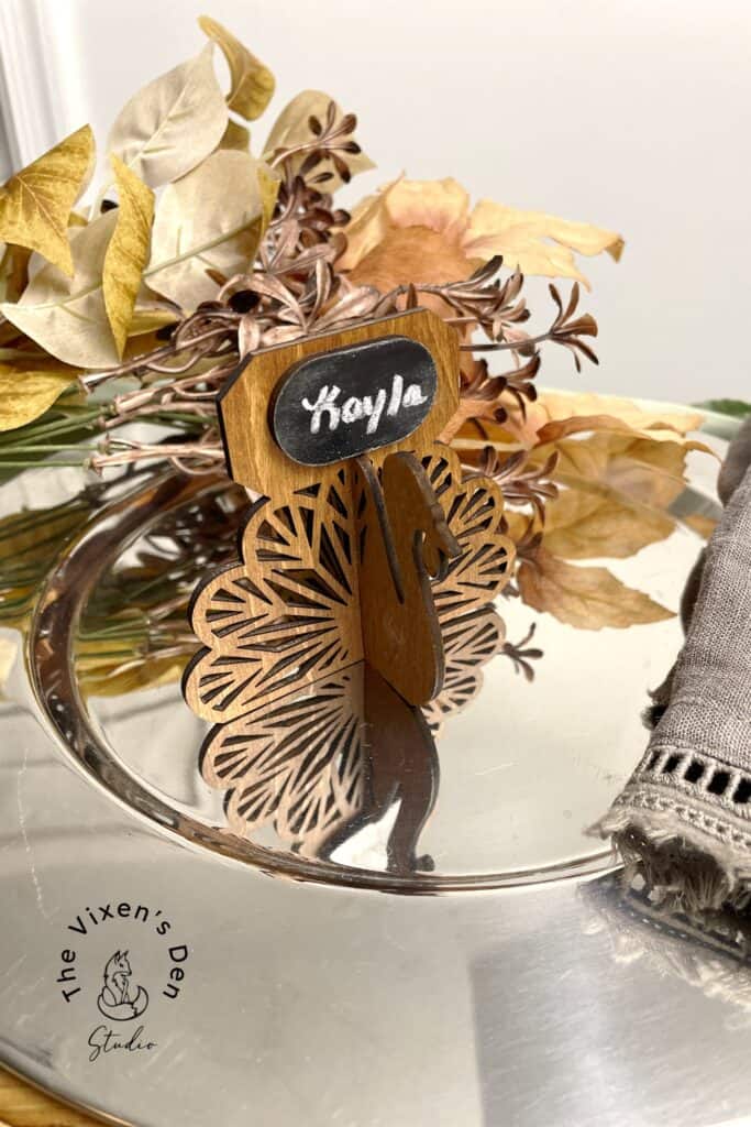 Wooden peacock-shaped place card holder with "Kayla" written on a chalkboard, placed on a metal tray with dried leaves and a folded gray napkin.