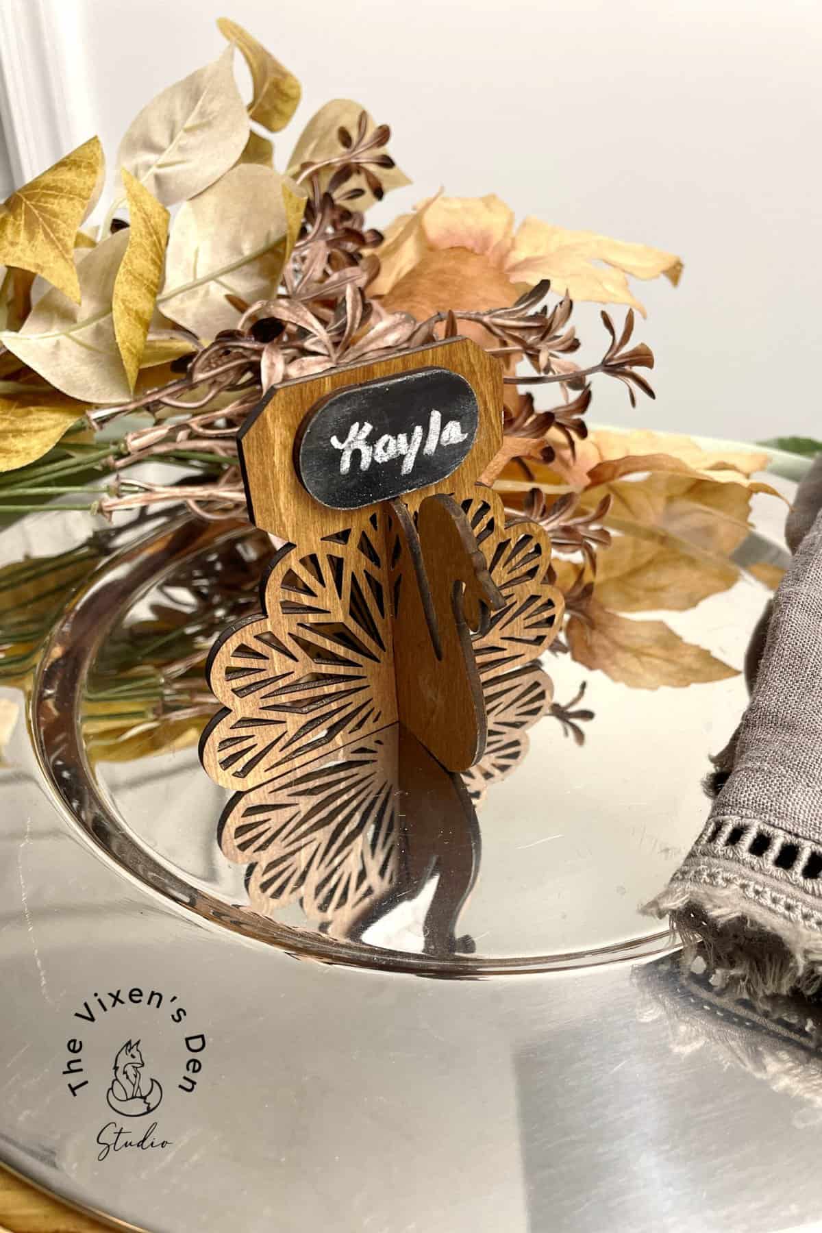 Wooden peacock-shaped place card holder with "Kayla" written on a chalkboard, placed on a metal tray with dried leaves and a folded gray napkin.