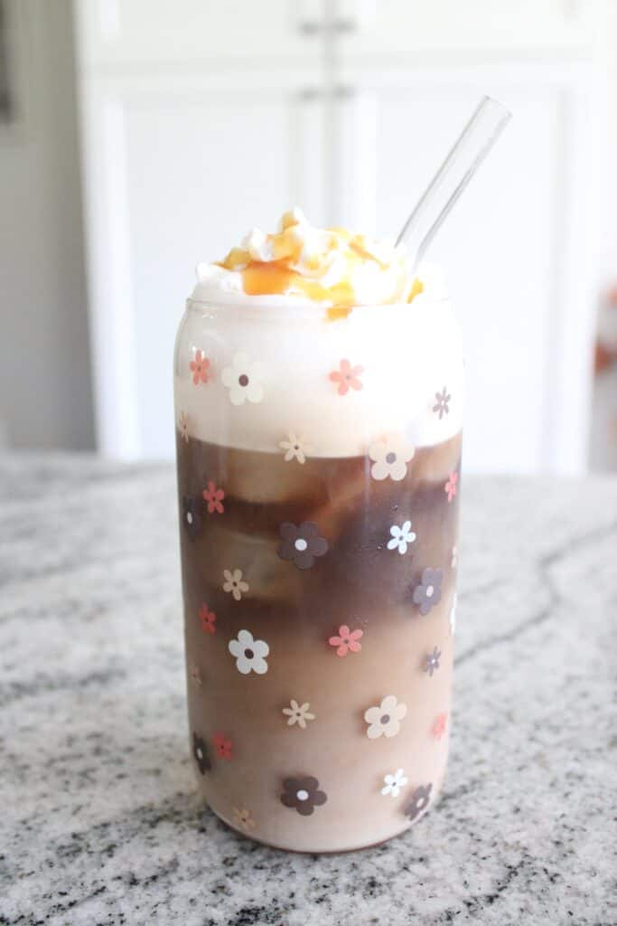 Iced coffee in a floral-patterned glass with whipped cream and caramel drizzle, placed on a marble countertop.