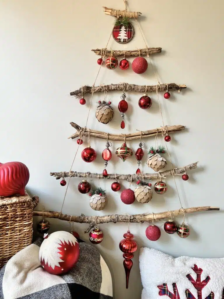 Wall-mounted Christmas tree made of twigs, adorned with red and gold ornaments, above a basket and chair with a decorative pillow.