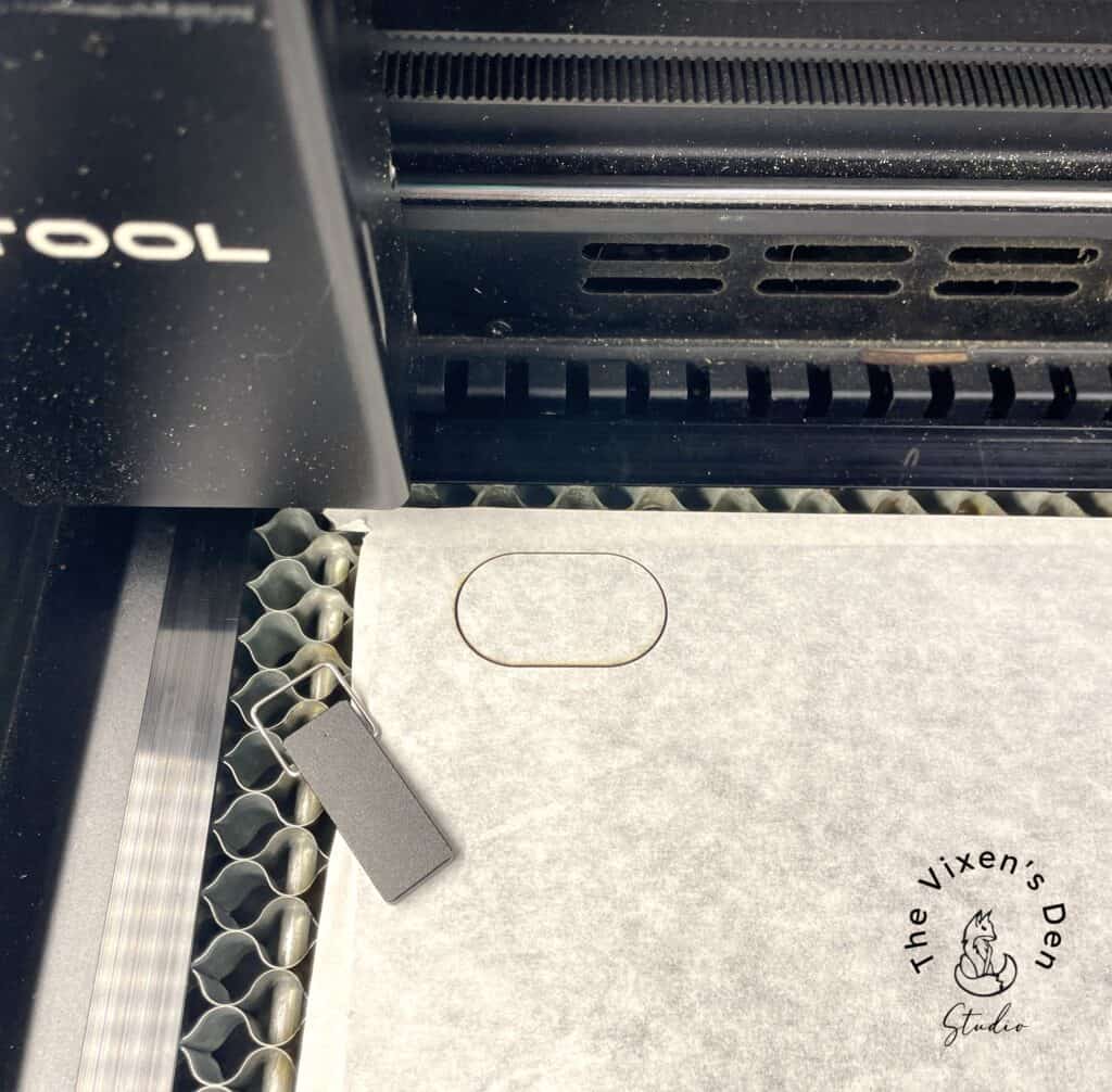 A laser cutter engraves a rectangular object on a sheet, with "The Vixen's Den" logo visible in the bottom right corner.