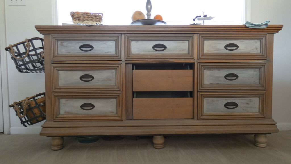 Wooden dresser with six closed drawers and one open middle drawer, showing two baskets on the left side. Items are on top, including a fruit bowl.