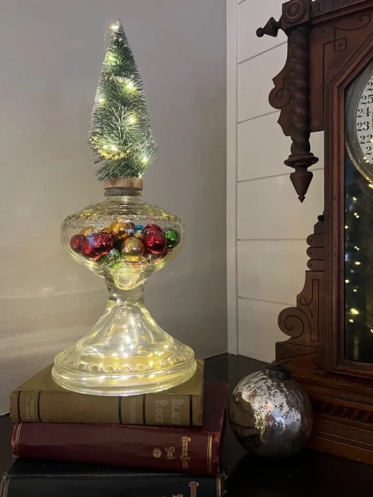 A glass lamp filled with colorful baubles is topped with a small, lit Christmas tree. It sits on a stack of books next to an ornate wooden clock.