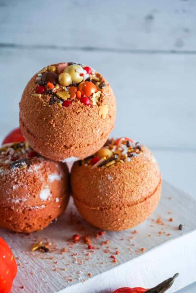 Three spherical bath bombs with colorful speckles are stacked on a white surface, with small decorative pumpkins nearby.