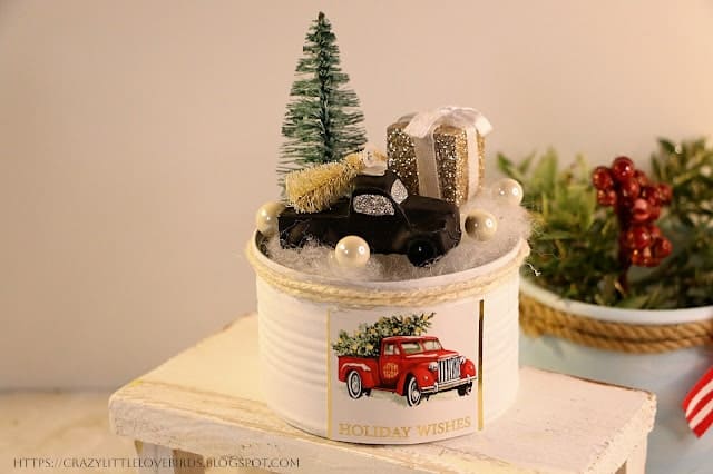 A festive decoration features a small black truck with a Christmas tree and gift on top, surrounded by faux snow and pearls, in a tin can labeled "Holiday Wishes," with a red truck illustration.