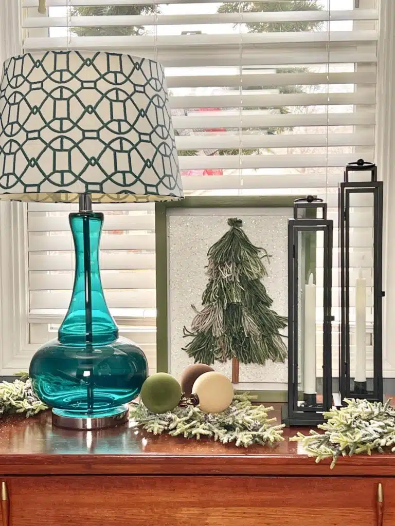 A teal lamp with a patterned shade sits on a wooden table beside decorative orbs, a framed tree, and two tall candle holders. White blinds cover the window in the background.