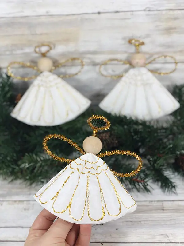 Hand holding a DIY angel ornament made with a wooden bead and gold pipe cleaners, with two similar ornaments and greenery in the background.