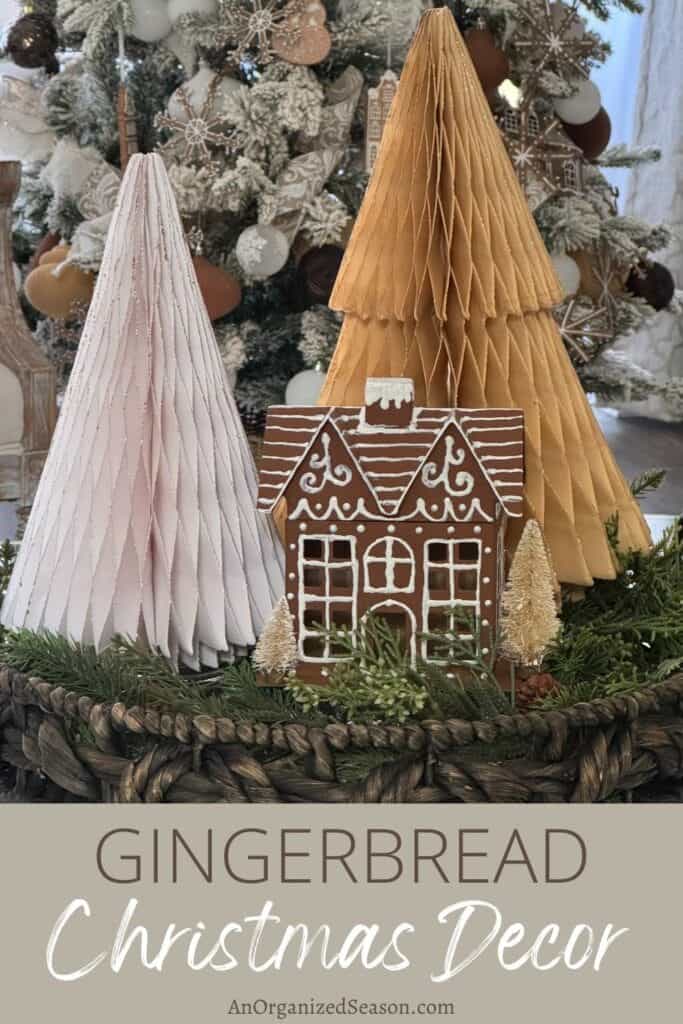 A gingerbread house is displayed between two large, textured cone trees, one white and one brown, set among evergreen branches with small white ornaments in the background.