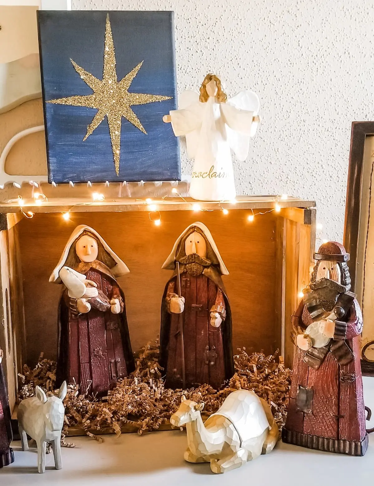 Nativity scene with figurines of Mary, Joseph, and a shepherd inside a wooden box. Animals are placed in front, and a star decoration and angel figure are displayed behind.