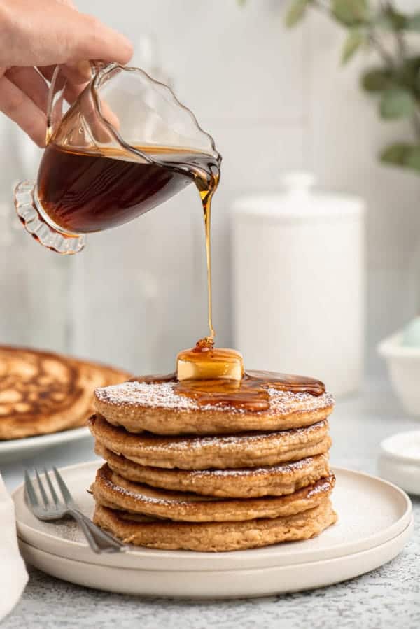 A hand pours syrup onto a stack of pancakes topped with butter on a plate, with a fork beside it.