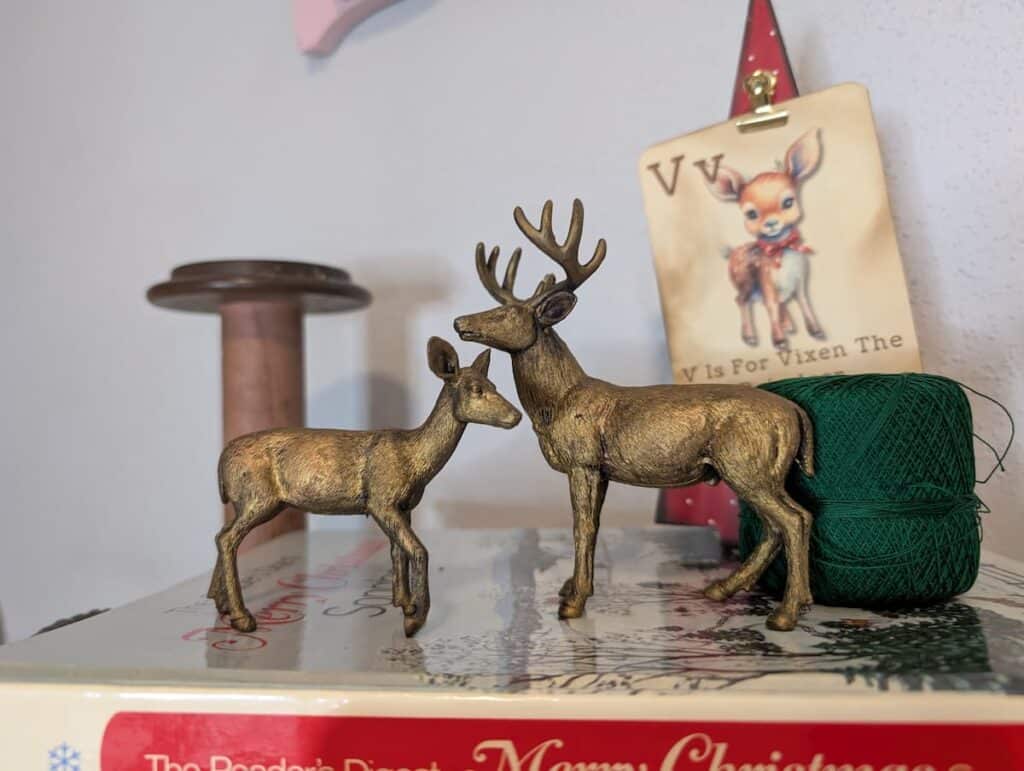 Two brass deer figurines on a surface with a spool of green thread and a book. A small reindeer artwork on the wall in the background.