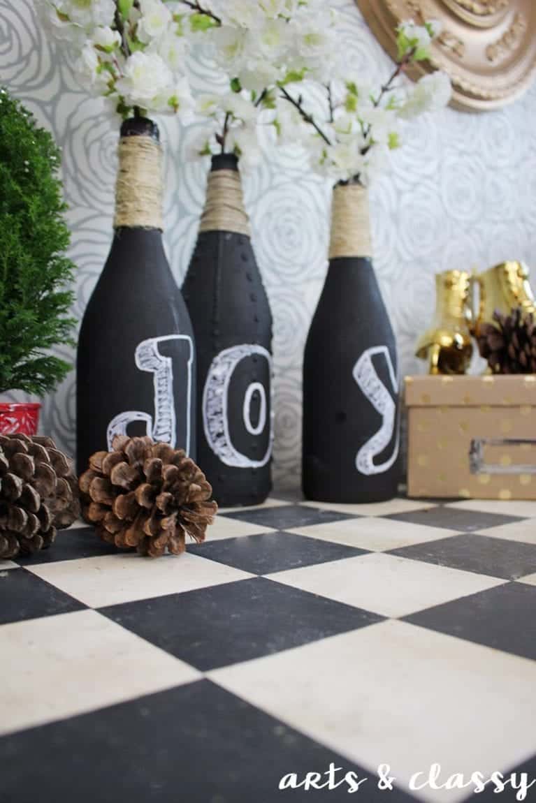 Three black bottles with "JOY" spelled out in white sit on a checkered surface, decorated with twine and white flowers. Pinecones and small gold reindeer figurines are nearby.