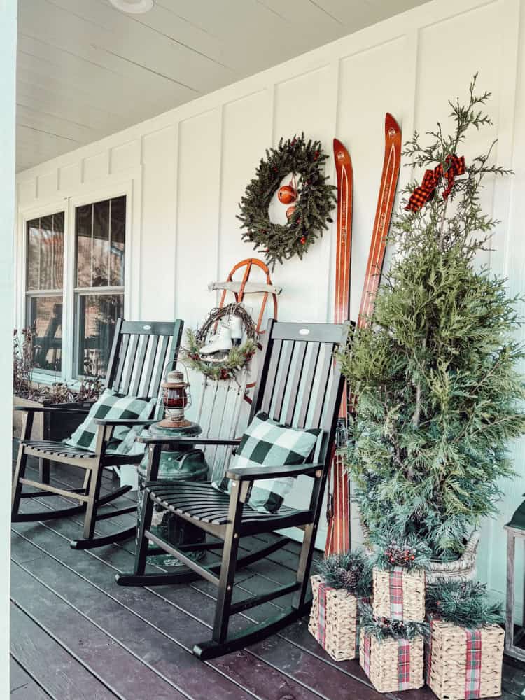 A porch with two black rocking chairs, green plaid cushions, wooden skis, a sled, and holiday decorations, including a wreath and gift boxes, arranged under a covered roof.