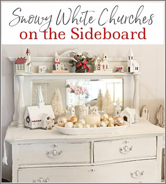 A white sideboard displays a collection of small snow-covered church models, decorative trees, and a bowl of gold and silver ornaments.