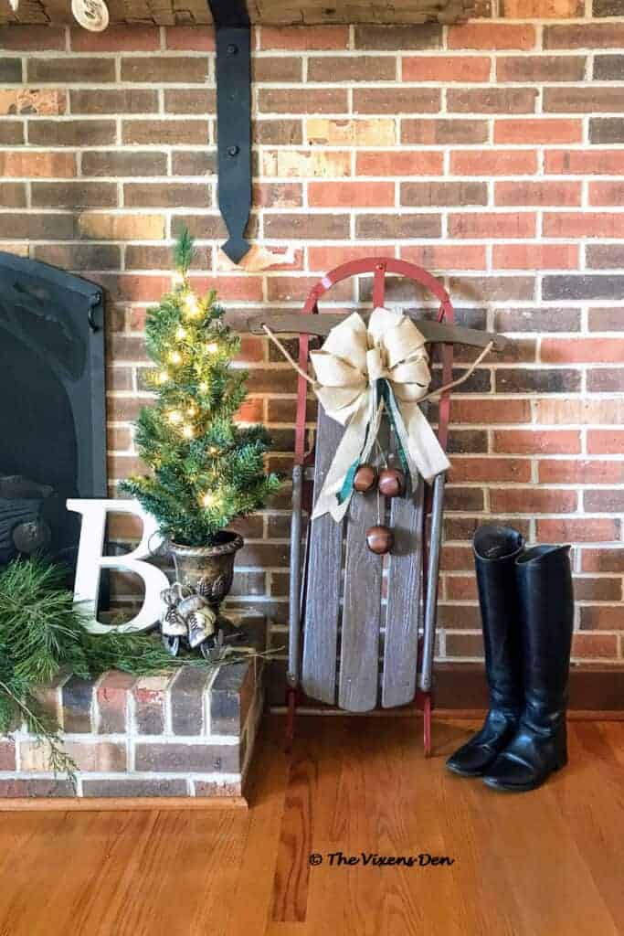 Vintage sled with a bow leans against a brick fireplace, next to a small decorated tree and black boots.